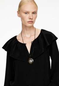 Blonde Frau mit einer schwarzen Bluse mit gerafftem Ausschnitt und einer runden Schildpatt-Halskette an einem schwarzen Band.