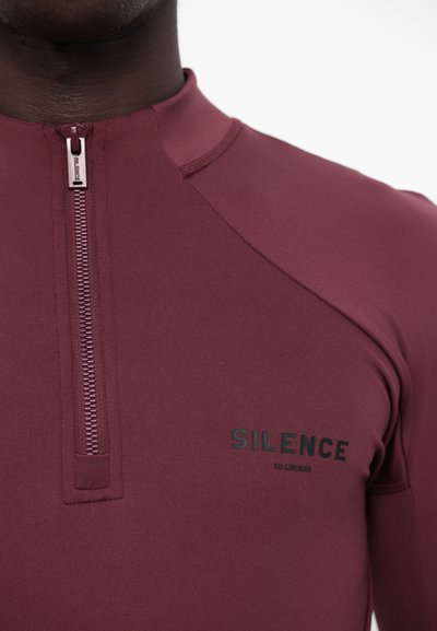 Maroon long-sleeve top with a high collar, featuring a front zip and “SILENCE” logo in black on the chest. Smooth, stretchy fabric.