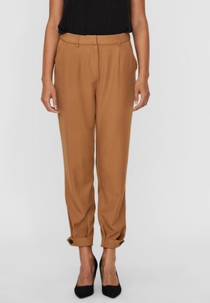 Frau trägt hoch taillierte, hellbraune, verjüngte Hose mit umgeschlagenen Säumen und schwarzen Knöpfen, kombiniert mit schwarzen spitz zulaufenden Schuhen und einem schwarzen Oberteil.