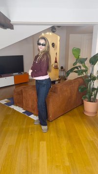 Pruun fliisist diivan ümmarguse kujuga, tumesinised teksad, roosa särk ja mustad päikeseprillid. Hele puidust põrand, potis roheline taim.