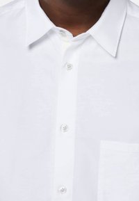 Närbild av en persons överkropp som bär en skarp vit skjorta med krage och en enda bröstficka, som visar knappar och tygtextur.