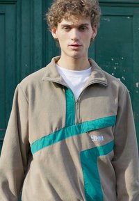 Sudadera de polar beige con una franja diagonal turquesa y cuello con cremallera. Presenta un acabado texturizado y un pequeño logo en la parte delantera.