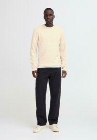 Beige sweater with a textured finish, crew neckline; paired with dark pants and sneakers. Simple, modern design, with neutral colors.