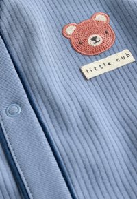 Hellblaue gerippte Strickjacke mit einer Stickerei eines braunen Bären und einem Etikett "kleiner Welpe". Sie hat einen runden Knopfverschluss und eine weiche Textur.