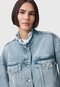 Chaqueta de mezclilla azul claro con cuello deshilachado, botones plateados y dos bolsillos frontales. Tejido texturizado con áreas desgastadas y un corte relajado.