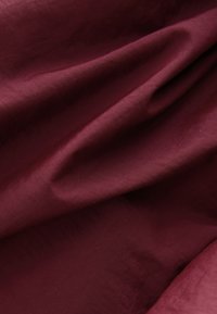 Tissu bordeaux profond et lisse avec des plis doux et une texture subtile, créant de légères ombres et des reflets délicats.