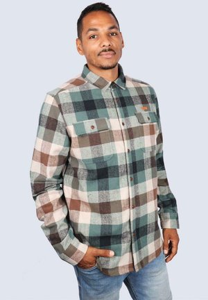 Man with short hair wearing a green, brown, and white plaid button-up shirt with chest pockets and blue jeans, standing with one hand in pocket.