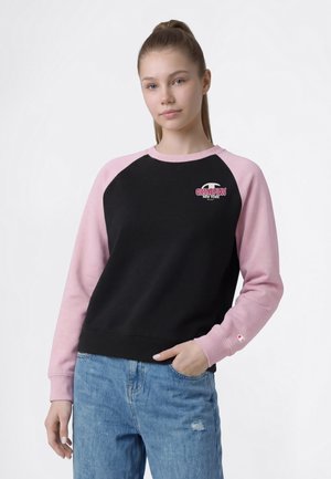 Ung kvinde med håret samlet bagpå iført en sort og pink Champion sweatshirt med lange ærmer og lyseblå jeans med rippet detalje.