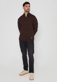 Pullover zip-up marrone scuro con maniche lunghe e colletto alto, abbinato a jeans neri e sneakers beige con suola chiara.
