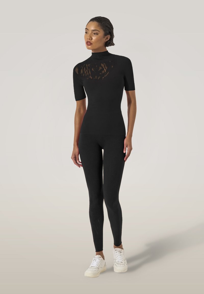 Svart åtsittande topp med korta ärmar och hög krage, med genomskinlig blommig spetsöverdrag över bröstet. Matchas med matchande leggings och vita sneakers.