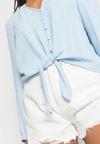 Ljust blå blus med lång ärm, knytning i midjan och knapppdetaljer. Matchad med vita denimshorts med fransad kant.