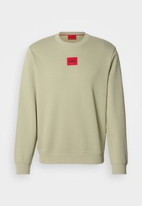 Sweatshirt beige à col rond avec poignets et ourlet côtelés, présentant un petit patch logo carré rouge avec "HUGO" centré sur la poitrine.
