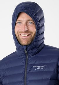 Marineblaue Steppjacke mit hohem, kapuzenförmigem Kragen, ausgestattet mit gesteppten Bereichen und einem Reißverschluss. Logo auf der Brust.