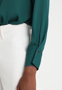 Blusa color azul verdoso con una textura suave, con mangas largas y un acento de corazón dorado en el puño. Combinada con pantalones blancos.