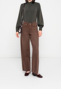 Jean en denim marron à jambes larges, assorti à un haut à manches longues vert foncé, avec un col montant et des manches bouffantes, ainsi que des chaussures foncées.