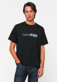 Mladý muž oblečený v čiernom tričku Calvin Klein a modrých džínsoch, postavený s jednou rukou v vrecku na pozadí bielej steny.