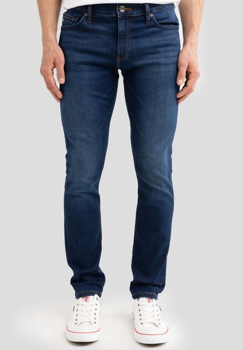 Mężczyzna ubrany w ciemnoniebieskie dżinsy typu slim-fit i białe trampki, stojący na tle jednolitego jasnoszarego tła.