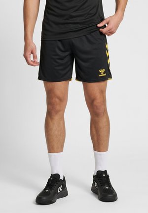 Una imagen recortada muestra a un modelo con pantalones cortos deportivos negros con chevrones amarillos de Hummel, calcetines blancos y zapatillas negras, levantándose el bajo de la camiseta.