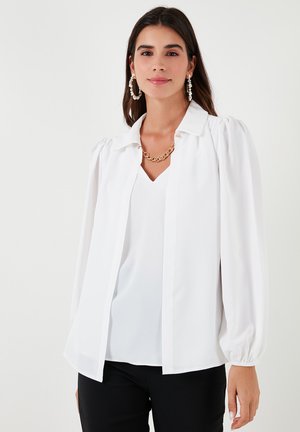 White blouse with a collared neckline and pleated puff sleeves, featuring a button closure. Smooth fabric with a relaxed fit, layered over a v-neck top.