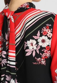 Blusa rossa e nera con motivo floreale, accenti a righe e dettaglio collo annodato. Presenta un tessuto liscio e colori vivaci.