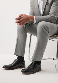 Schwarze Leder-Herrenschuhe mit Kappenspitz, Schnürsenkeln und brauner Kontrastsohle. Kombiniert mit einem grauen, maßgeschneiderten Anzug und schwarzen Socken.