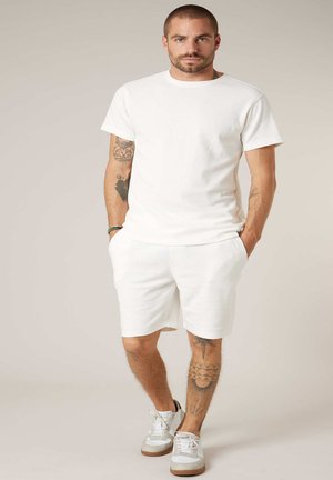 Homme tatoué portant un t-shirt blanc, un short blanc et des baskets blanches, debout les mains dans les poches devant un fond uni.