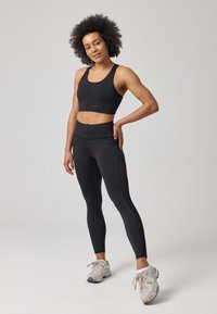 Soutien-gorge de sport noir et leggings avec empiècements en maille, design ajusté, texture lisse, et baskets grises neutres. Modèle debout avec une main sur la hanche.