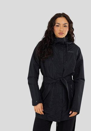 Donna con capelli lunghi e ricci indossa un impermeabile nero con cappuccio e cintura, dotato di tasche con zip, su sfondo grigio uniforme.