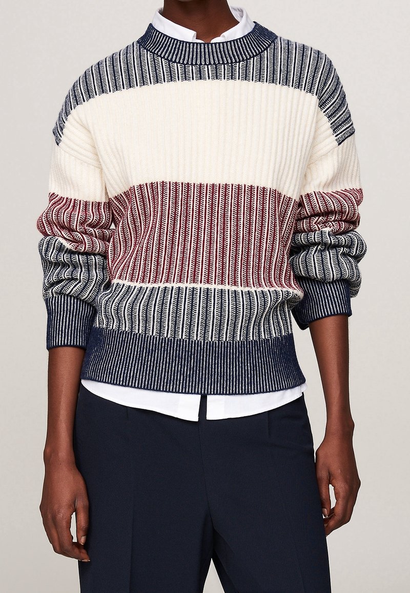 Personne portant un pull en maille côtelée avec des rayures horizontales bleu marine, blanc et rouge sur une chemise blanche à col et un pantalon bleu marine.