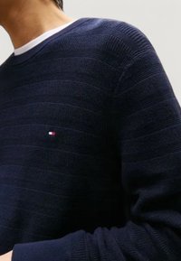 Man i en marinblå texturerad stickad tröja med en liten röd, vit och blå logotyp på bröstet över en vit t-shirt.