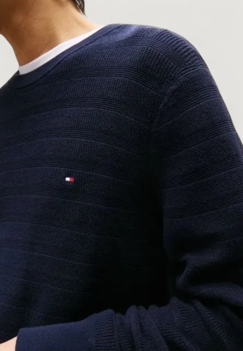 Man i en marinblå texturerad stickad tröja med en liten röd, vit och blå logotyp på bröstet över en vit t-shirt.