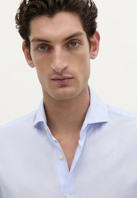 Camisa de botones azul claro con cuello puntiagudo, textura suave y un brillo sutil. Presenta un cierre de un botón en el cuello.