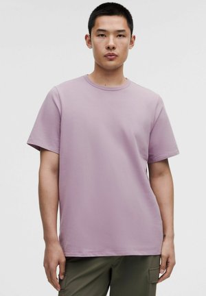 Jeune homme aux cheveux noirs courts portant un t-shirt lavande uni à manches courtes et un pantalon cargo vert olive, debout devant un fond blanc.