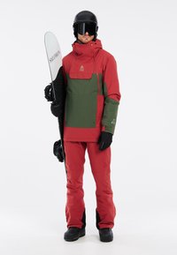 Rote und grüne isolierte Schneejacke mit einer Reißverschlusstasche, kombiniert mit roten Schneehosen und schwarzen Handschuhen. Hält ein Snowboard, trägt einen Helm und eine Skibrille.