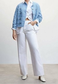 Cardigan bleu clair texturé avec boutons argentés porté sur une chemise blanche à boutons. Pantalon blanc à jambes larges et bottines blanches à bouts pointus.