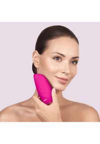Appareil de nettoyage facial en silicone rose avec un design incurvé, une surface texturée et deux boutons, tenu contre la joue d'une personne.