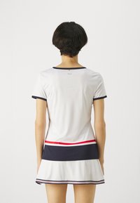 Bílé sportovní šaty s námořnickými a červenými pruhovanými akcenty, krátkými rukávy a volánkovou sukní. Materiál se zdá být hladký a lehký.