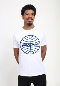 Bílý bavlněný tričko s námořnický modrou kruhovou nálepkou s textem "PAN AM" uprostřed, krátké rukávy a kulatý výstřih.