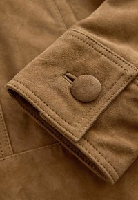 Colletto della giacca in suede con un bottone tondo e dettagli di cucitura. Il tessuto è di un marrone chiaro con una finitura morbida e testurizzata.