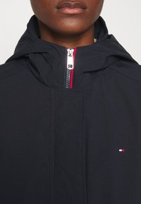 Námořnická modrá bunda s kapucí vyrobená z hladkého materiálu, s předním zipem a červenými a bílými akcenty, a malým logem na hrudi.