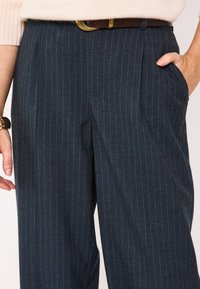 Pantalon sur mesure bleu marine à fines rayures, coupe ajustée, doté de poches latérales et d'une ceinture marron visible avec des boucles dorées.