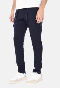 Joggers cargo blu navy con più tasche laterali, vita elastica e cordino. Abbinati a sneakers bianche e grigie su uno sfondo chiaro.