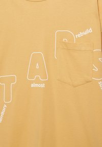 Tissu jaune avec une petite poche poitrine et de grandes lettres blanches contourées partiellement visibles, comprenant "almost", "rebuild" et "territory".