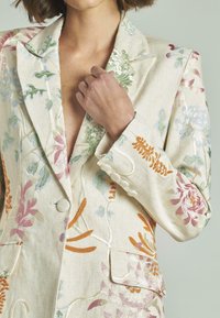 Blazer bordado en tela crema, con patrones florales en colores pastel. Detalles notables incluyen un cuello de chal y acentos de botón.