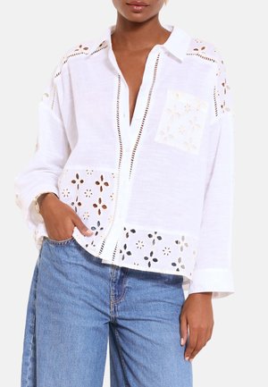 Kvinde iført løs hvid skjorte med knapper og blomsterudskæringsmønstre samt lyseblå højtaljede jeans, hånd i lommen.