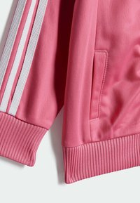 Casaco atlético rosa com punhos e bainha canelados, bolsos planos e riscas brancas nas laterais em tecido suave.
