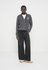 Γκρι ριμπ πουλόβερ με φερμουάρ και λογότυπο, σε συνδυασμό με μαύρο τζιν φαρδιάς γραμμής και λευκά αθλητικά παπούτσια με ταμπά εξωτερική σόλα.