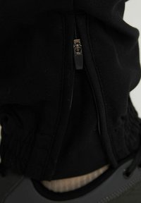 Vêtement en tissu noir avec une fermeture zippée, des accents élastiques en bas et une texture visible. Il présente un mélange de matériaux lisses et doux.