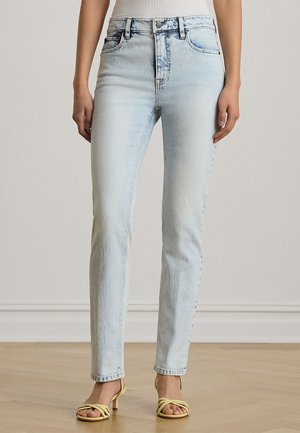 Femme portant un jean skinny taille haute bleu clair et des sandales à talons jaunes à lanières, debout sur un sol en bois devant un mur uni.