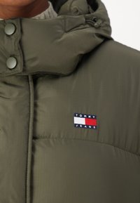 Olivgrön vadderad jacka med hög krage, tryckknappar och en Tommy Jeans-logotyp i blått, rött och vitt framtill.